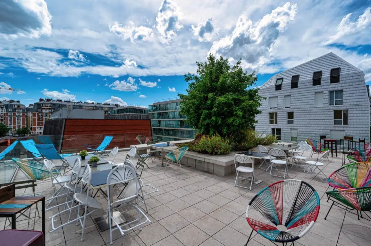 Terrasse ensoleillée avec chaises colorées et tables, idéale pour la maison de la conversation. Vue sur des bâtiments modernes et un ciel bleu.