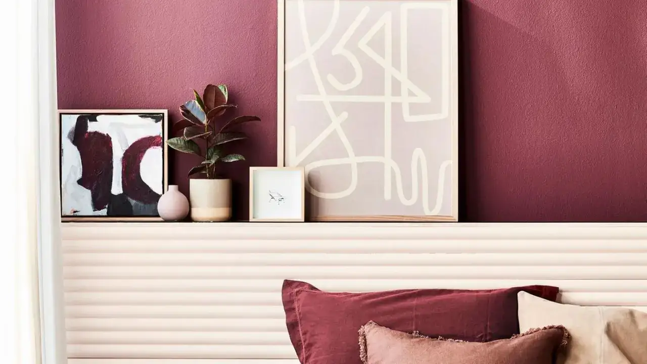 Ein Schlafzimmer mit Wand in englisches rosenrot, dekoriert mit Kunstwerken und einer Pflanze. Kissen in Rosatönen liegen auf dem Bett.