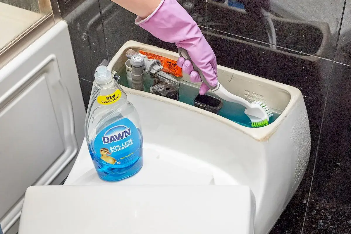 A gloved hand scrubs inside a toilet tank with a brush, next to a bottle of Dawn dish soap. This shows how to clean toilet tank.