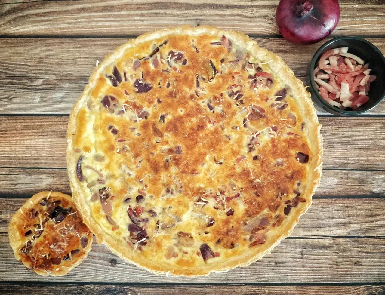 Une tarte à l'oignon dorée et appétissante, accompagnée d'une mini-tartelette, d'un oignon rouge et de lardons. Parfait pour un repas savoureux.