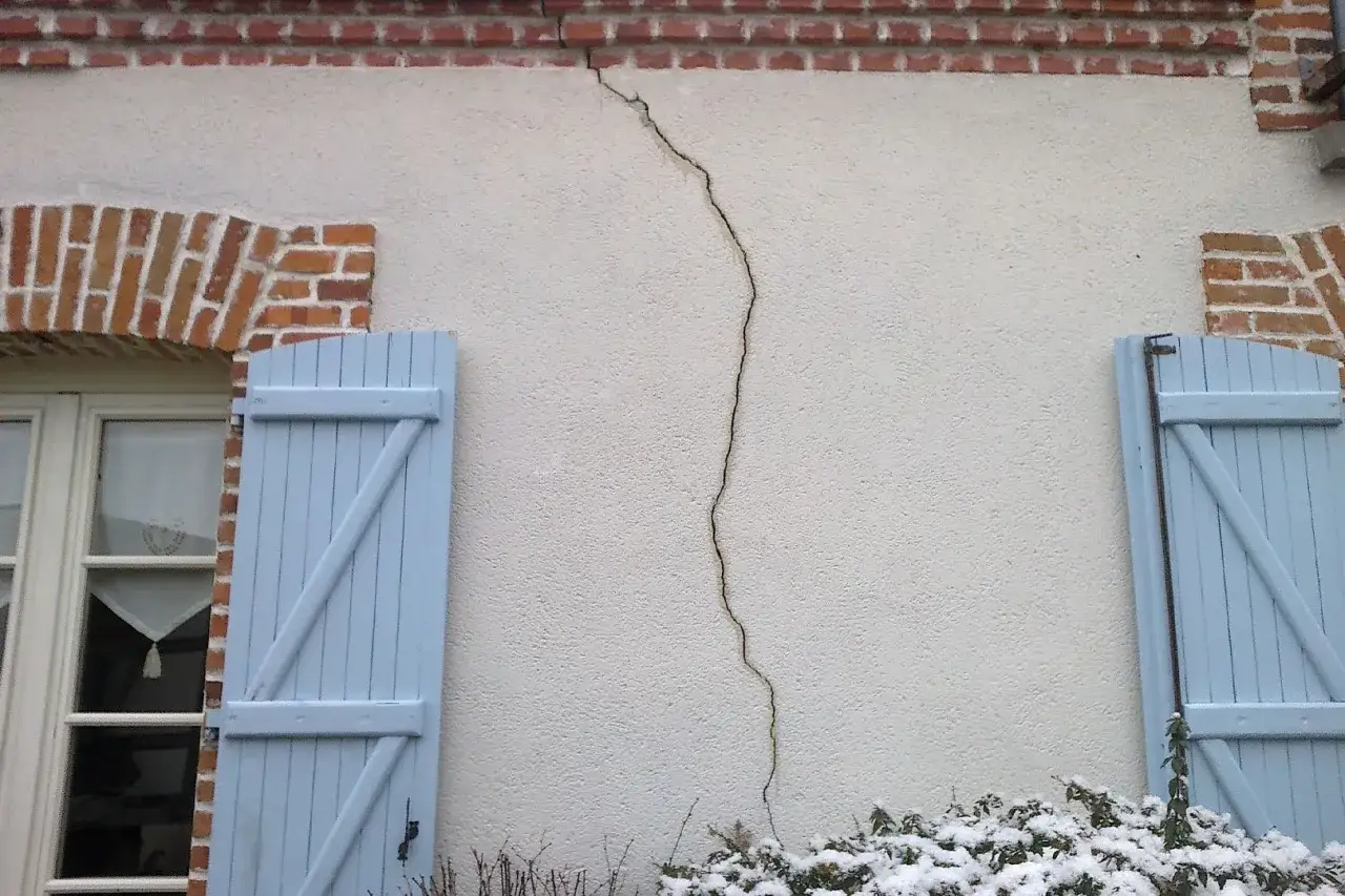 Une fissure traverse le crépi d'une maison, passant sous la corniche en briques et au-dessus d'un volet bleu.