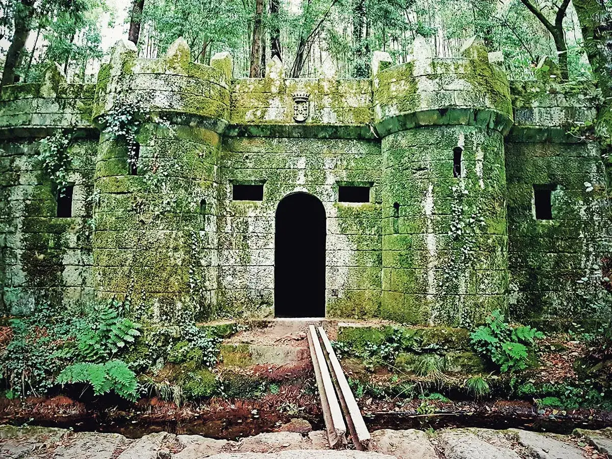 Antigua fortaleza cubierta de musgo en el bosque encantado de Aldán, con una entrada oscura y vegetación exuberante.