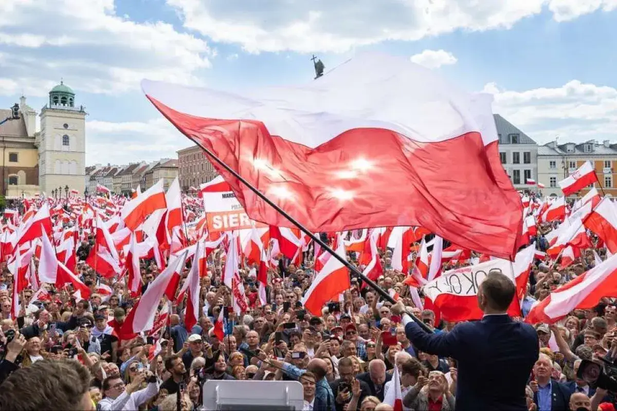 Tłum świętuje dzień flagi, unosząc setki biało-czerwonych flag. W centrum wielka flaga Polski, a w tle historyczna zabudowa.