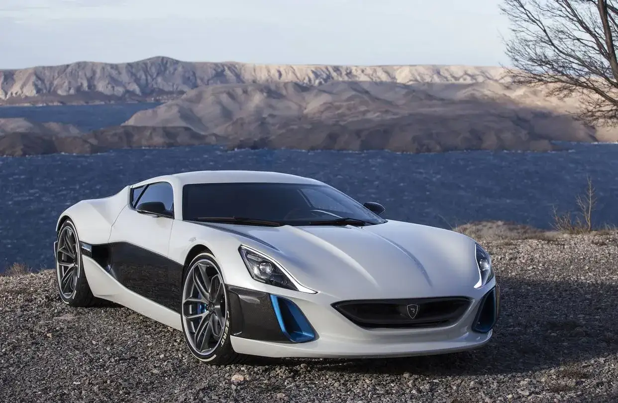 Un'auto elettrica sportiva bianca e nera, con dettagli blu, parcheggiata su un terreno ghiaioso con un lago e montagne sullo sfondo.