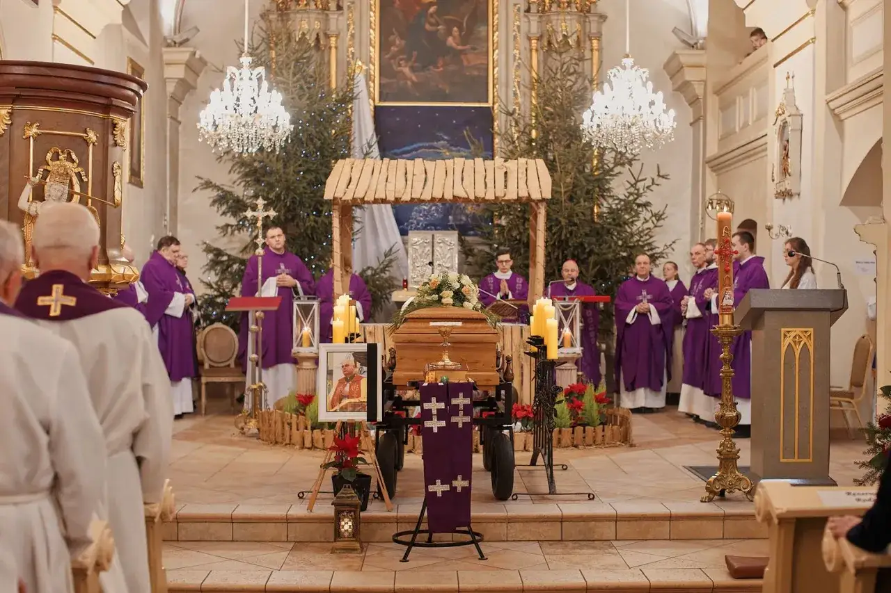 Ksiądz wygłasza mowę pogrzebową przy trumnie w kościele. Wokół zgromadzeni duchowni w purpurowych szatach.