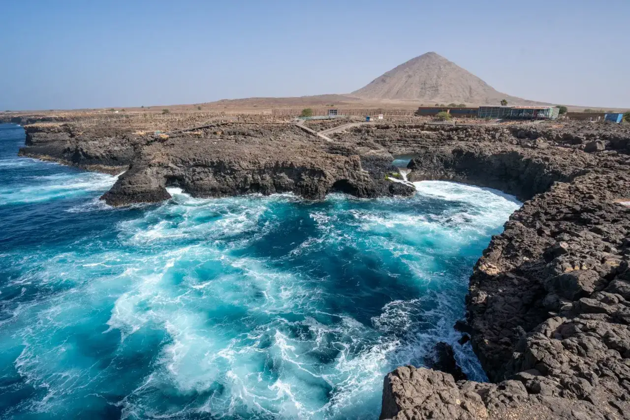 Türkisblaues Meer brandet an felsige Küsten auf den Kapverden im Juli. Im Hintergrund ragt ein vulkanischer Berg auf.