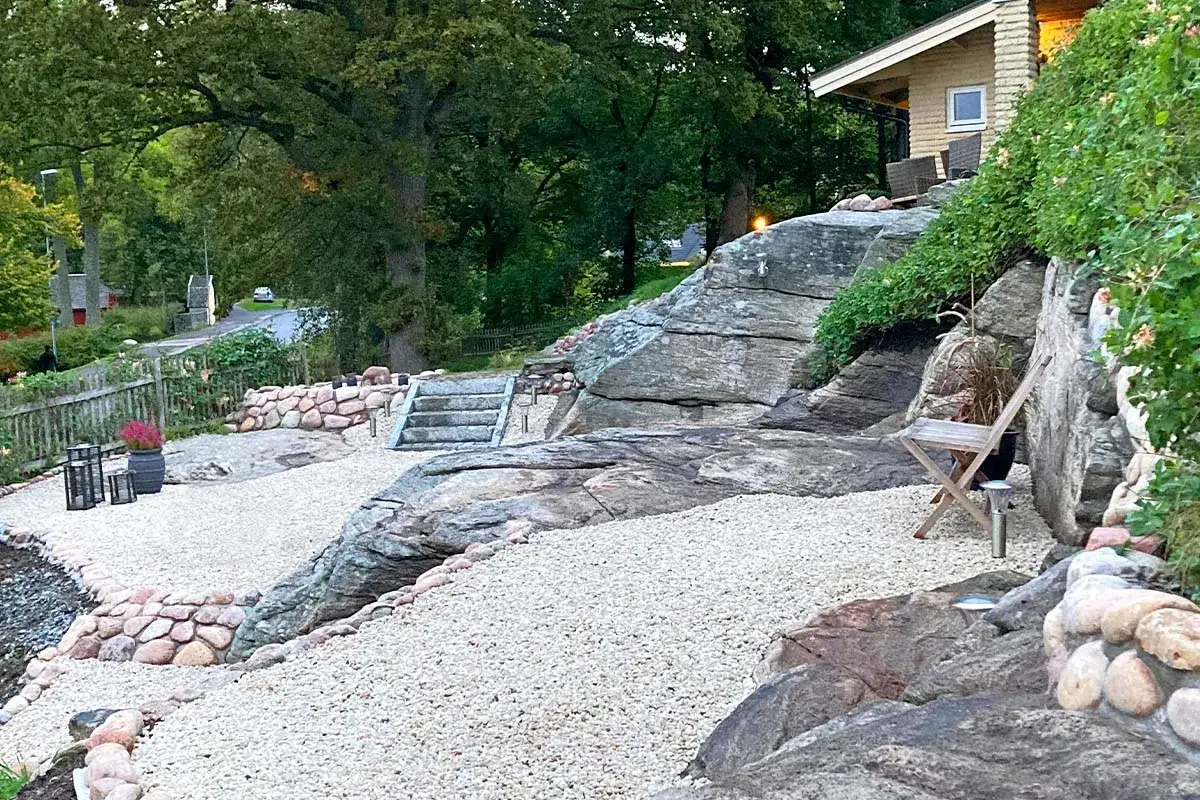 En vacker uteplats sten med grusg&aring;ngar, stenmurar och en trappa som leder upp mot ett hus. En stol st&aring;r redo f&ouml;r avkoppling.