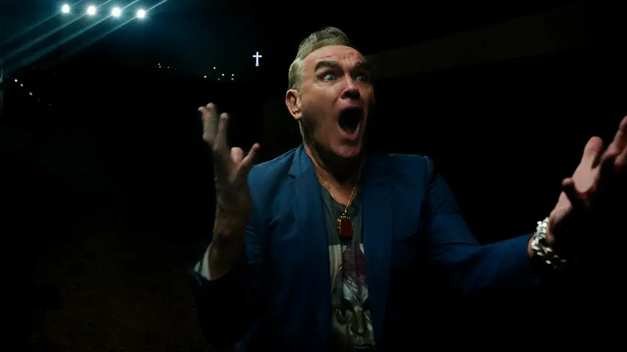 Morrissey en Zaragoza, con gesto dramático y brazos extendidos, bajo luces de escenario y una cruz iluminada.