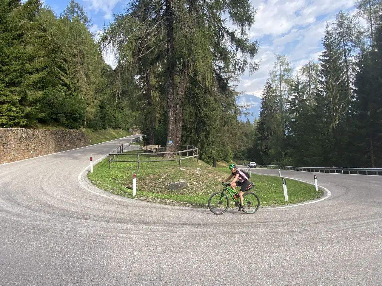 Ciclista su strada tortuosa nel **campo Carlo Magno**, circondato da alberi e montagne.