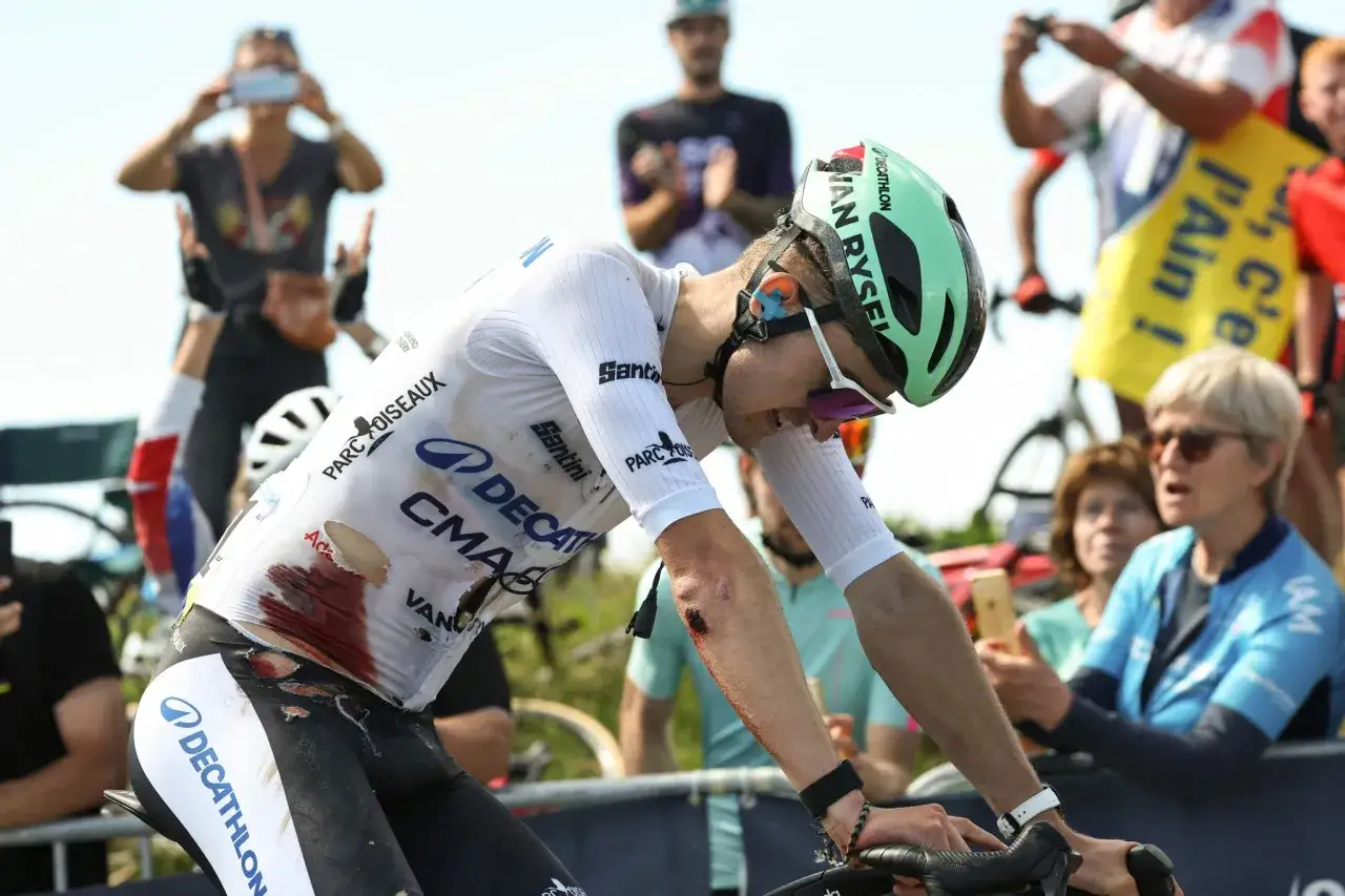 A cyclist in a torn white jersey, showing injuries, rides during the Tour de France. Spectators watch.