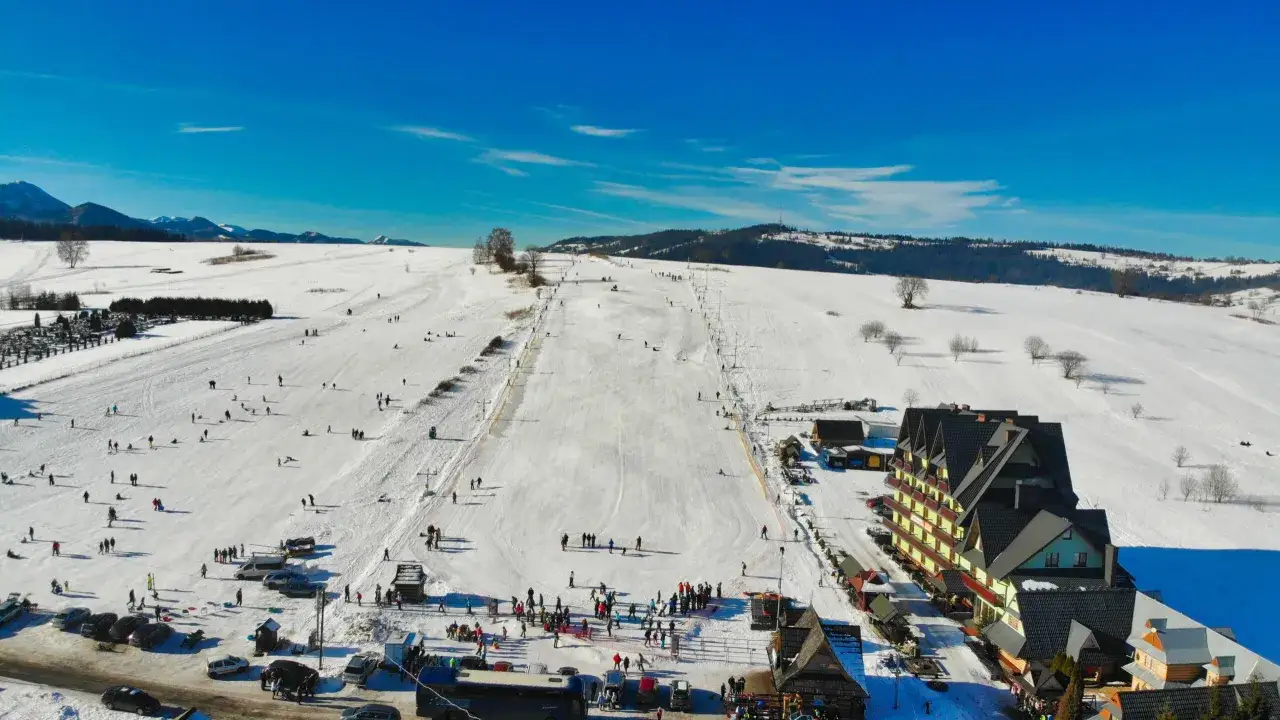 Słoneczny dzień na stoku, gdzie działa wyciąg w Zakopanem. Ludzie jeżdżą na nartach i snowboardach, a w tle widać g&oacute;ry.