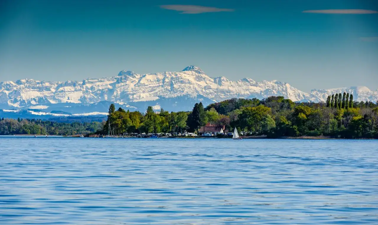 Ein idyllischer August-Urlaub am See mit schneebedeckten Bergen im Hintergrund. Ein Segelboot gleitet &uuml;ber das ruhige Wasser.