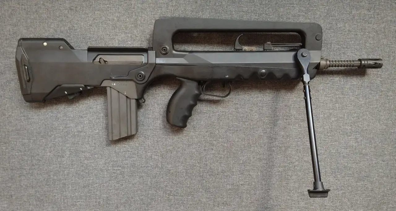 Czarny karabin FAMAS z charakterystycznym uchwytem i dwójnogiem, leżący na szarym tle.