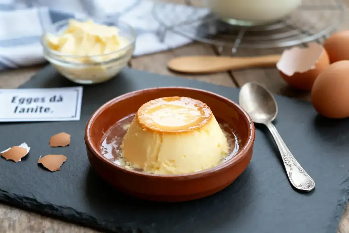 Un d&eacute;licieux oeuf au lait, onctueux et dor&eacute;, servi dans un ramequin en terre cuite. Des coquilles d'oeufs et une cuill&egrave;re compl&egrave;tent la sc&egrave;ne.
