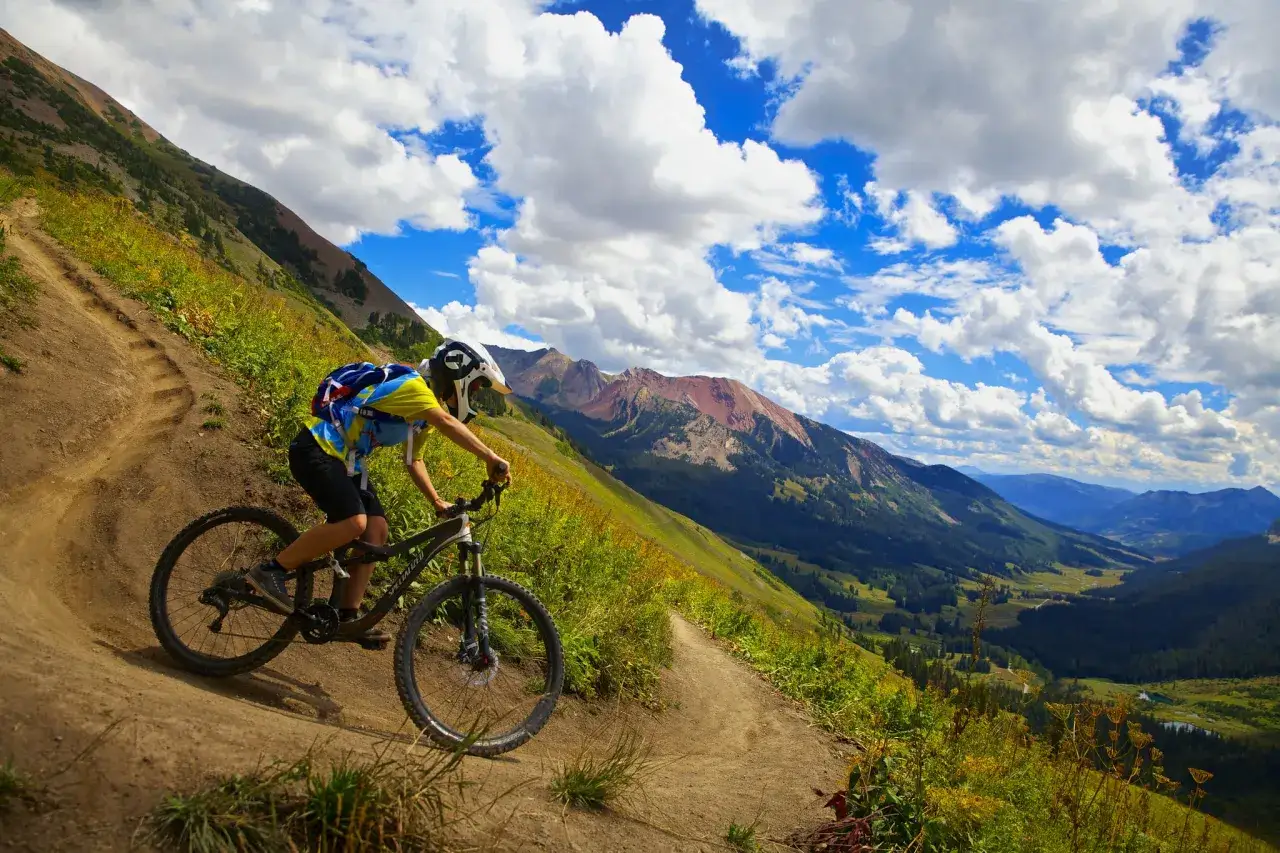 A mountain biker navigates a dusty trail on a steep slope, experiencing the best mountain biking in Colorado with stunning mountain vistas and a dramatic sky.