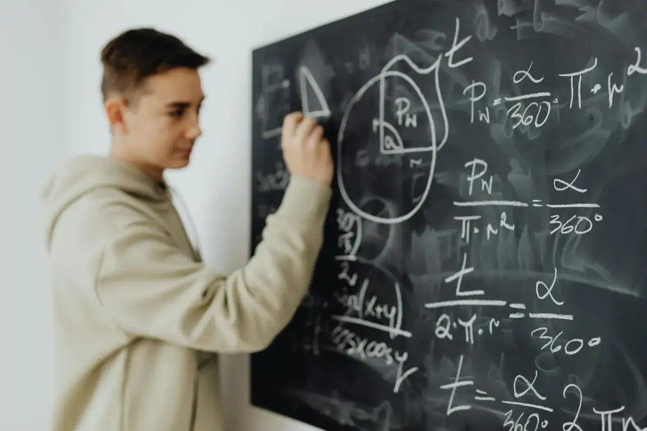 Chłopak w bluzie rozwiązuje zadania matematyczne na tablicy. To obrazek dla uczniów wybierających kierunki liceum.