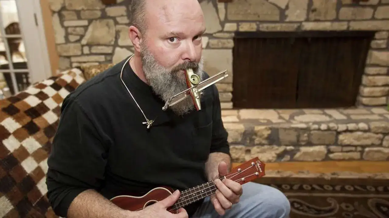 Homme barbu jouant du ukulélé et du kazoo musique, assis près d'une cheminée en pierre.