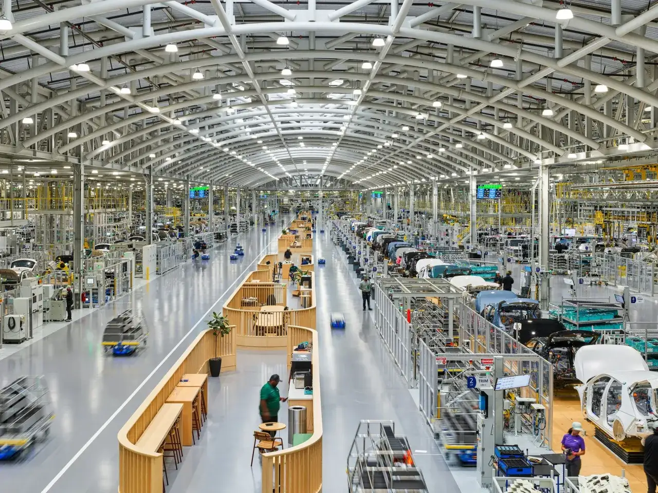 Stor bilfabrik där bilar tillverkas. Automatiska robotar och arbetare rör sig längs produktionslinjen.