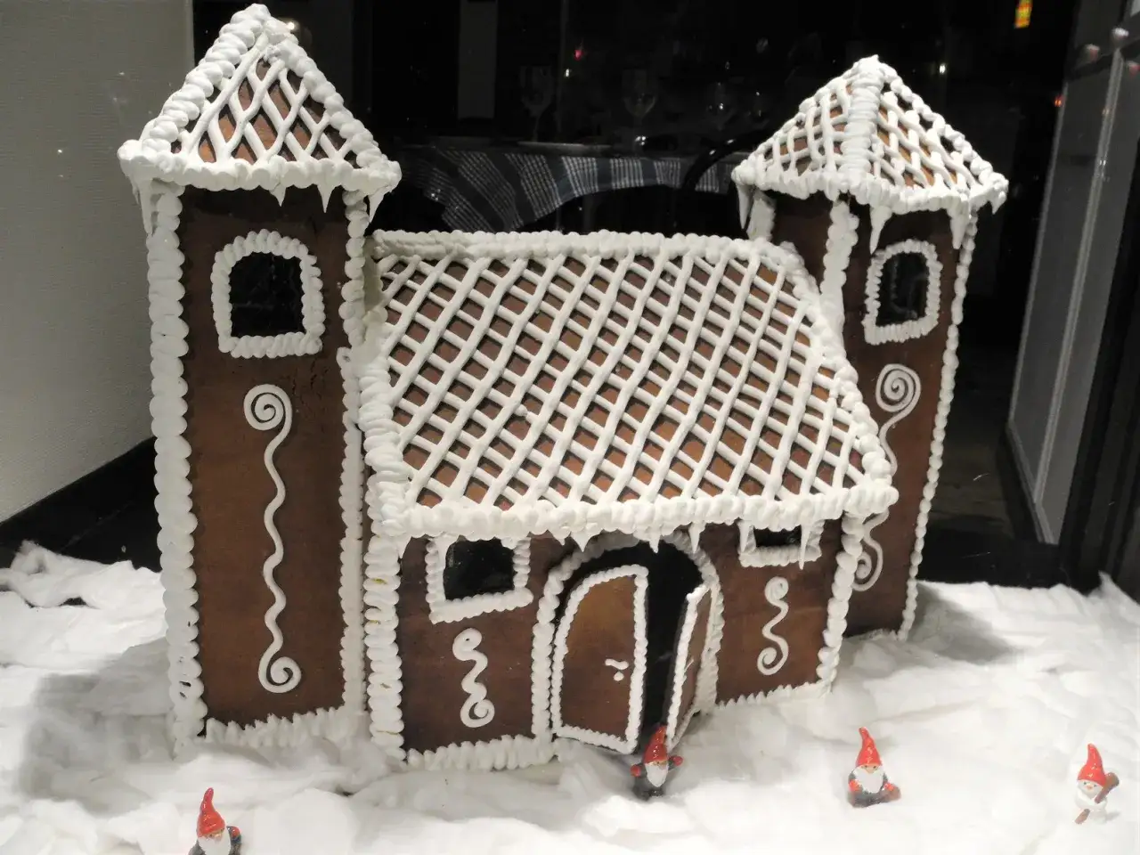 Ett imponerande pepparkakshus med detaljerad kristyr, som en liten borg i snön. Små tomtar tittar fram.