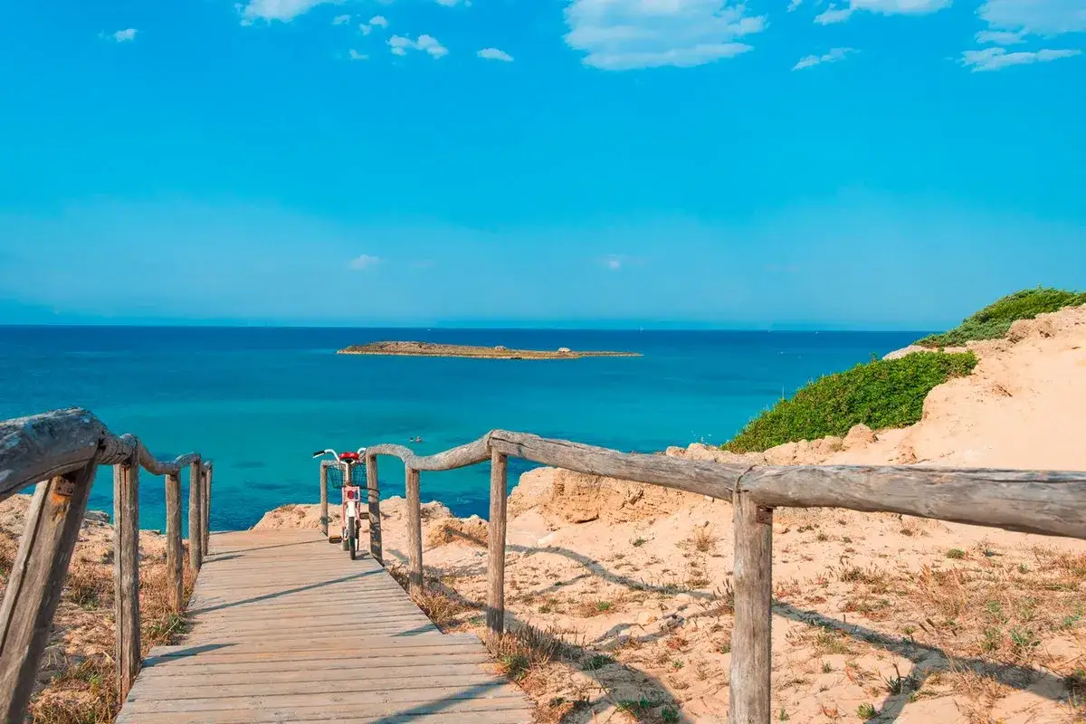 Sentiero in legno verso il mare cristallino, con un'isola in lontananza. Questo è il panorama mozzafiato della marina di Ugento, dove si trova questo angolo di paradiso.