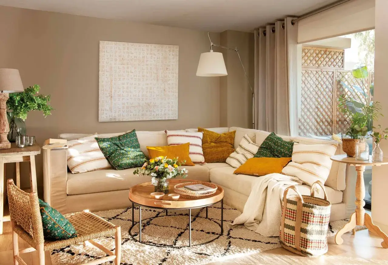 Sofá chaise longue beige con cojines coloridos, alfombra de pelo largo con diseño geométrico, mesa de centro redonda de madera y cesta de mimbre.