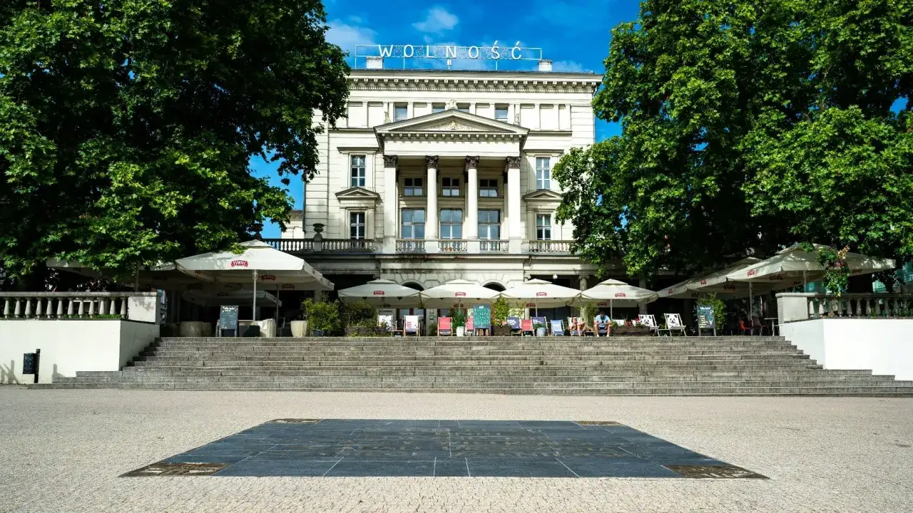 Plac Wolności w Poznaniu z monumentalnym budynkiem i kawiarnianymi ogródkami pod parasolami.