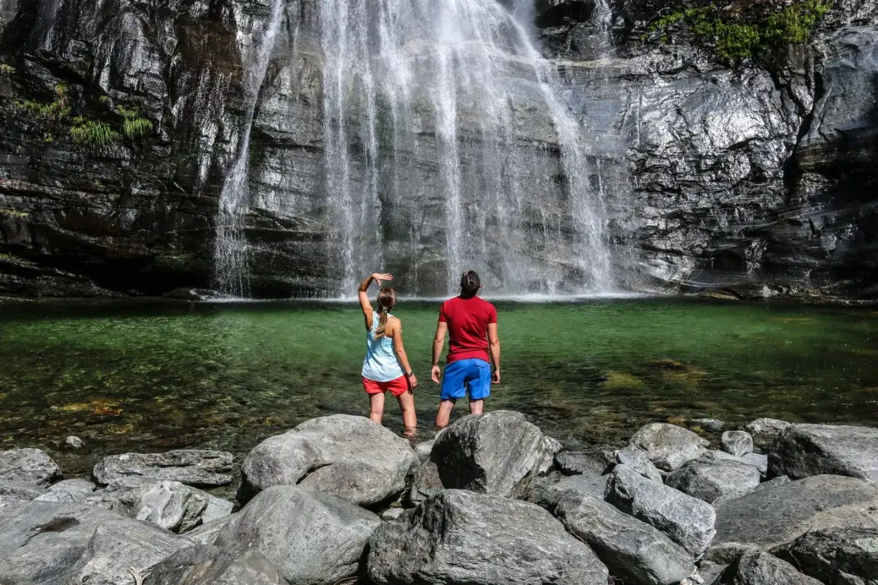 Zwei Wanderer stehen im klaren Wasser vor einem tosenden Wasserfall im Valle Maggia.