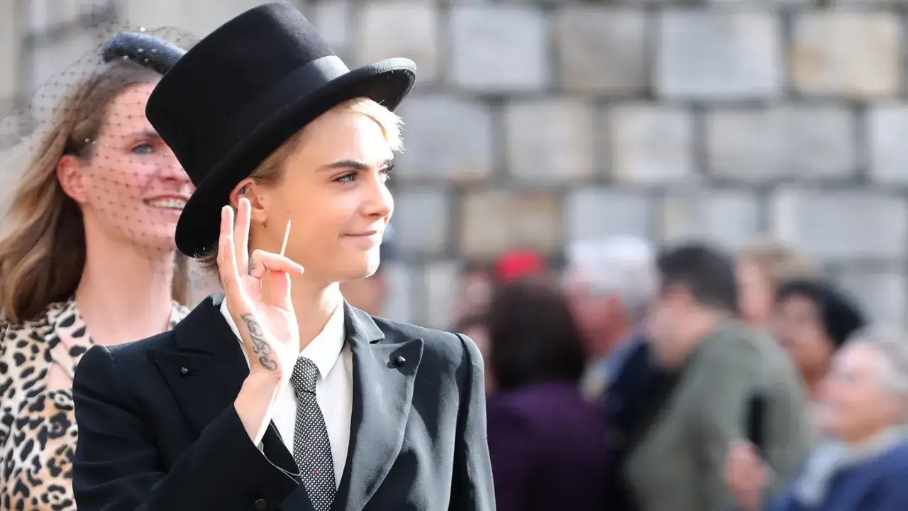 Una tomboy que es, con sombrero de copa y traje, saluda con la mano.