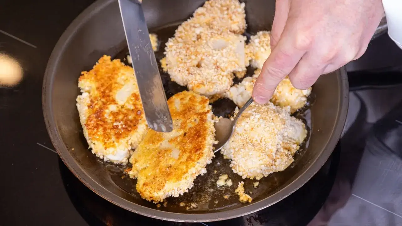 Goldbraune Blumenkohl-Schnitzel brutzeln in der Pfanne. Ein Messer und ein L&ouml;ffel helfen beim Wenden.