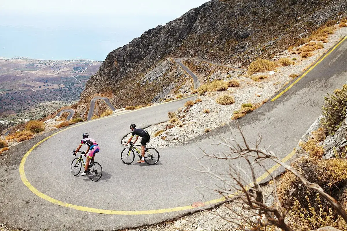 Zwei Radfahrer meistern Serpentinen auf Kreta. Ein Rennrad-Abenteuer mit atemberaubender Aussicht auf das Meer und die zerklüftete Landschaft.