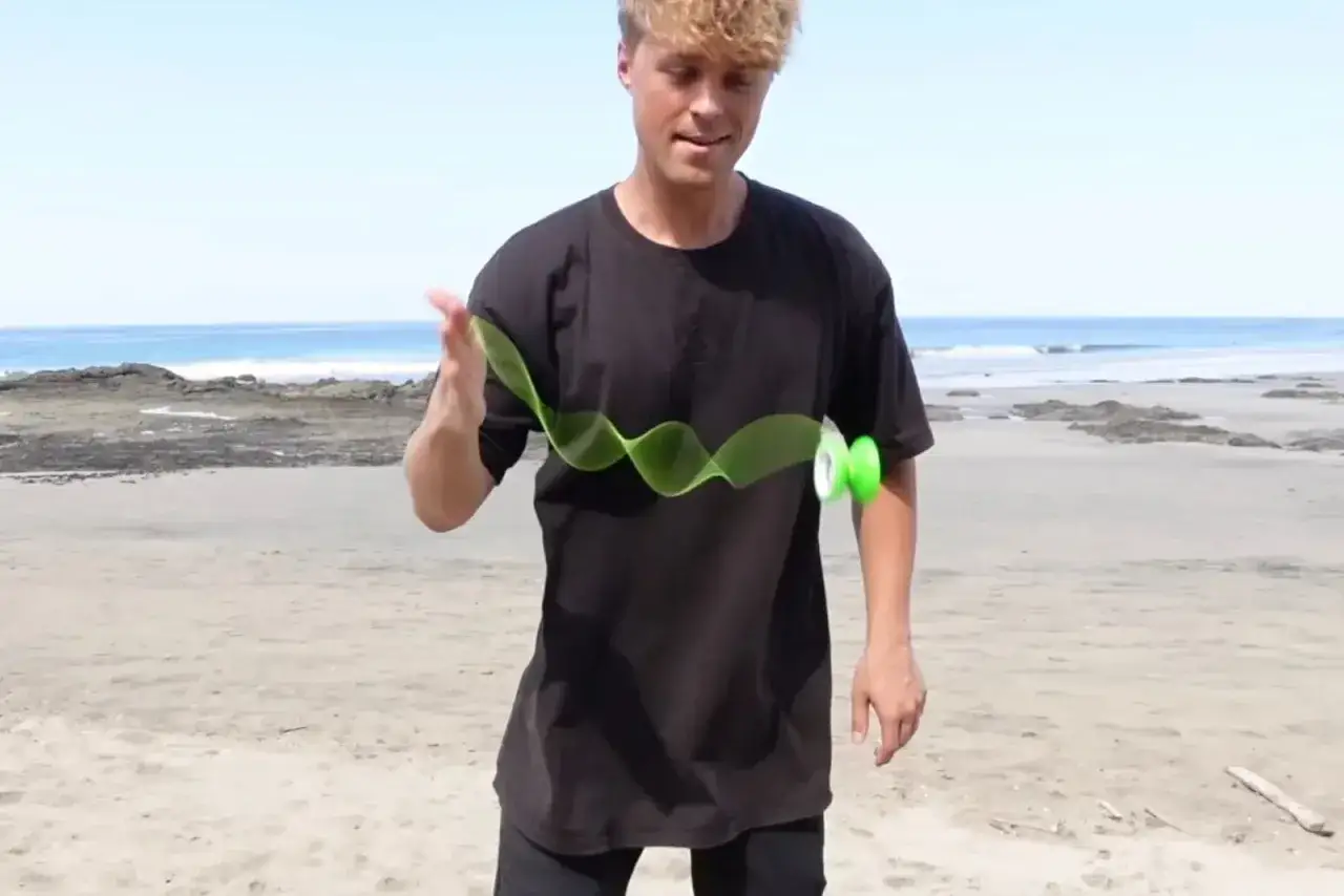 En ung man med blont hår leker med ett grönt yoyo på en strand.