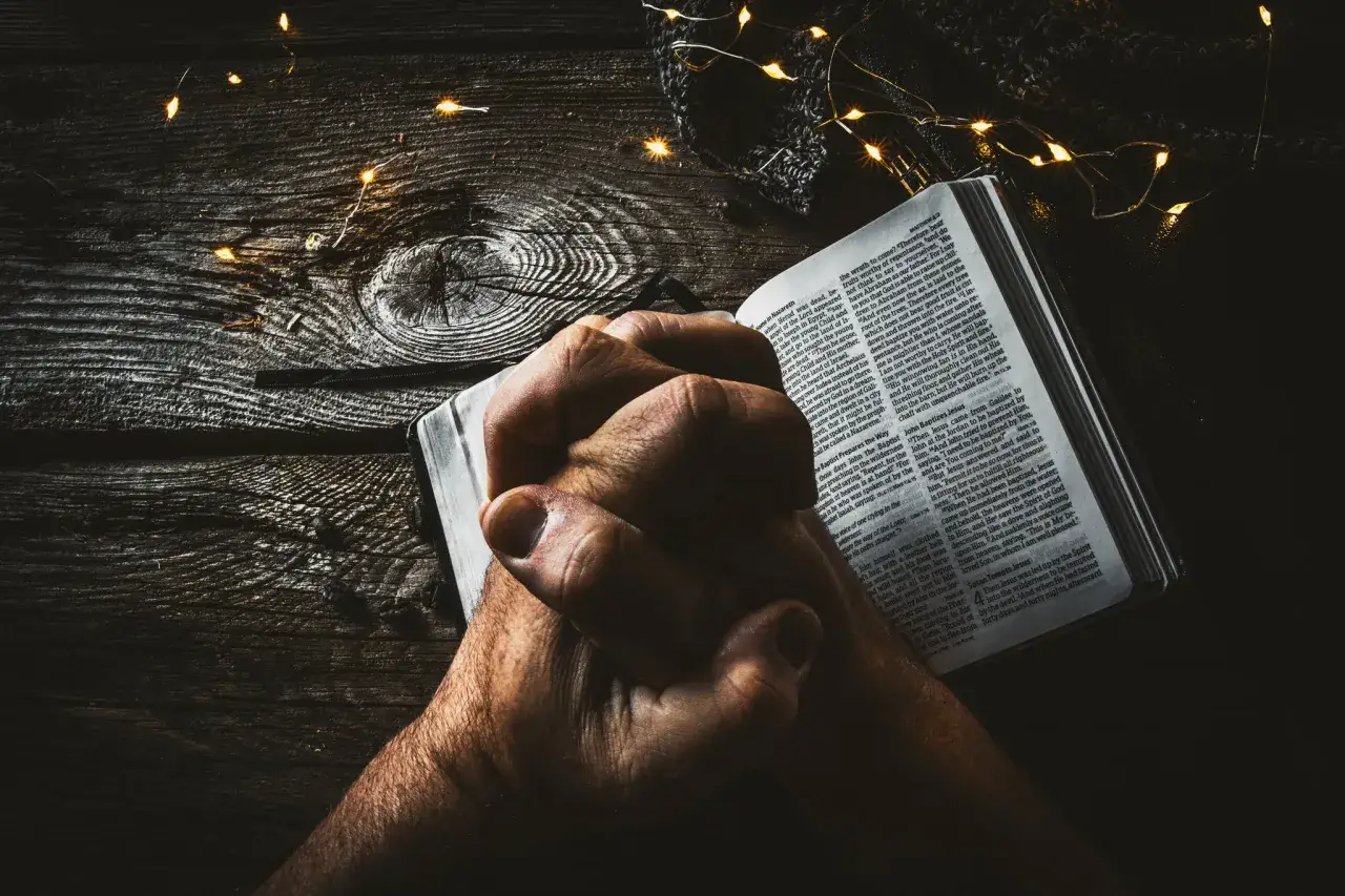 Hands clasped in prayer over an open Bible, illuminated by fairy lights, signifying moments of Christian prayer.