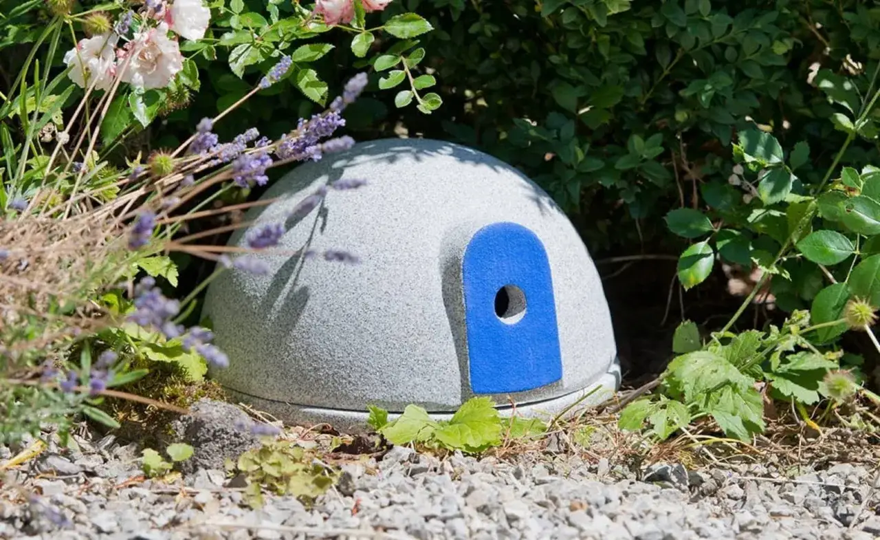 Ett runt, gr&aring;tt hus med bl&aring; d&ouml;rr&ouml;ppning, perfekt f&ouml;r humlor bon, omgivet av lavendel och gr&ouml;nska.