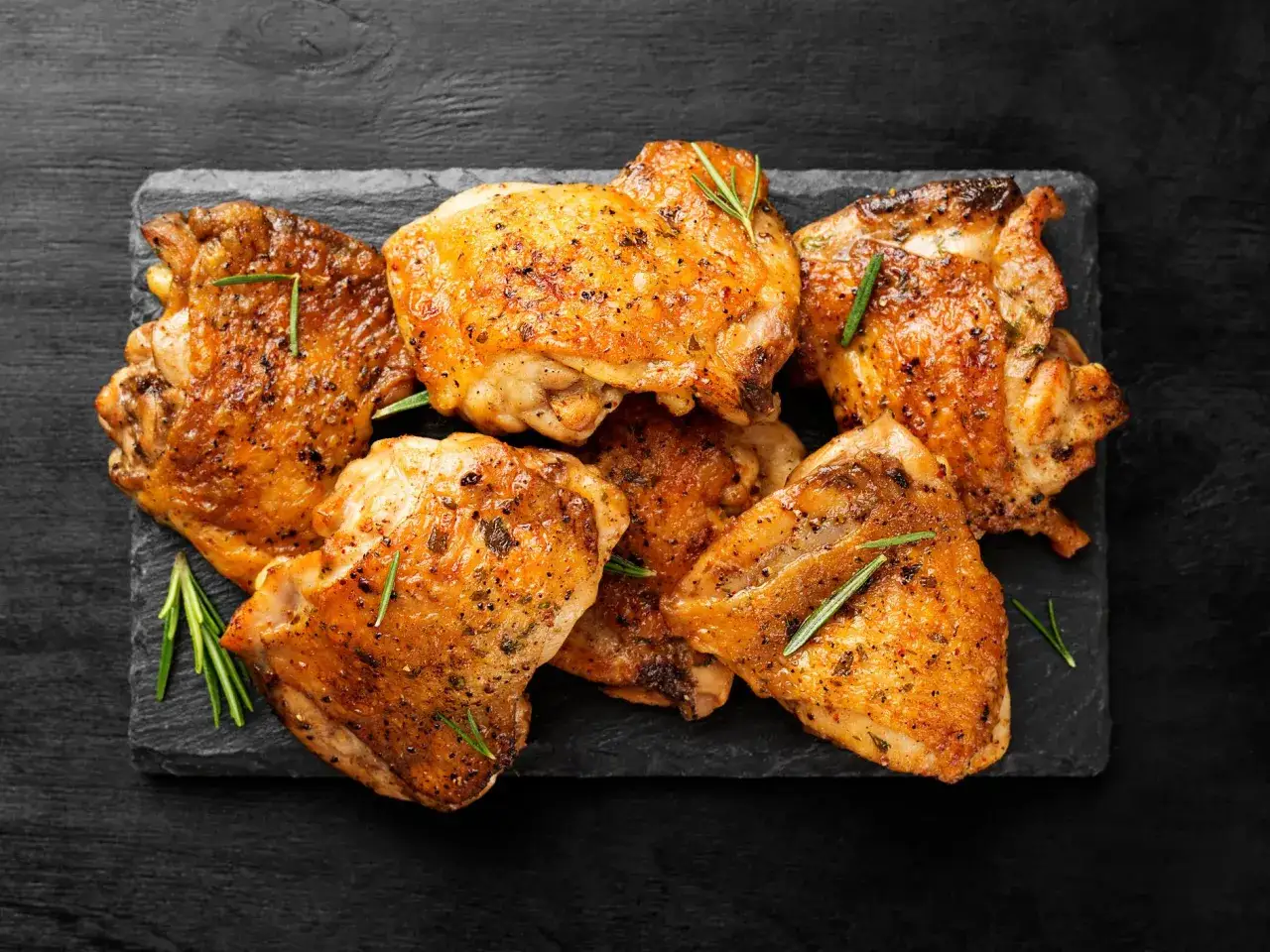 Plusieurs hauts de cuisse de poulet dorés et croustillants, assaisonnés d'herbes, disposés sur une planche d'ardoise.