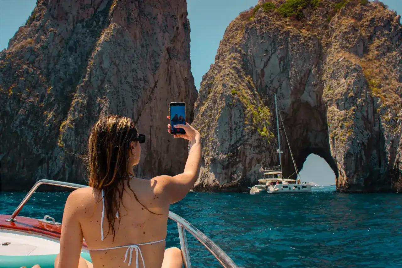 Donna in barca riprende con lo smartphone l'arco roccioso che indica dove si trova la Grotta Azzurra, con catamarano e altre imbarcazioni.