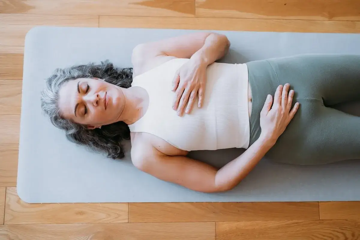 Frau in entspannter Pose, H&auml;nde auf Brust und Bauch, genie&szlig;t eine **Meditation im Liegen** auf einer Matte.