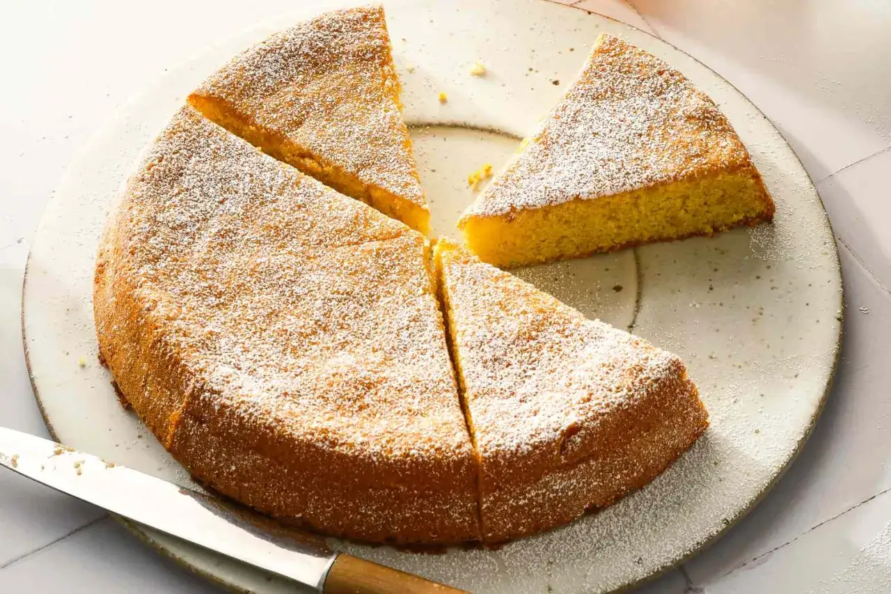 Ein saftiger zitronenkuchen ohne mehl, best&auml;ubt mit Puderzucker, liegt auf einem Teller. Ein Messer liegt daneben.