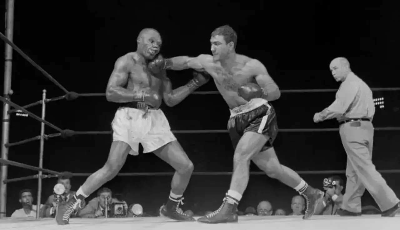 Rocky Marciano reaches for a punch during a boxing match, with an opponent and referee in the ring.