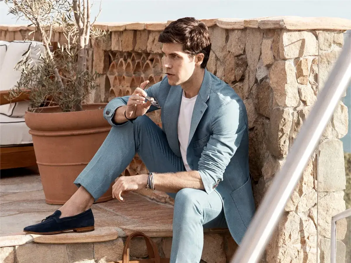 Hombre con traje azul claro y mocasines oscuros, sentado en un patio de piedra con una planta grande.