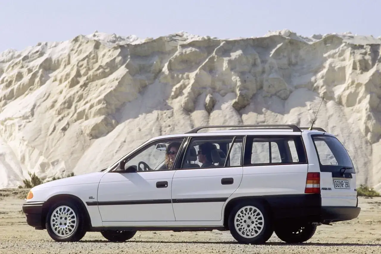 Biały Opel Astra Classic 1999 kombi z dwoma osobami w środku, zaparkowany na pustkowiu z piaskowymi wzgórzami w tle.