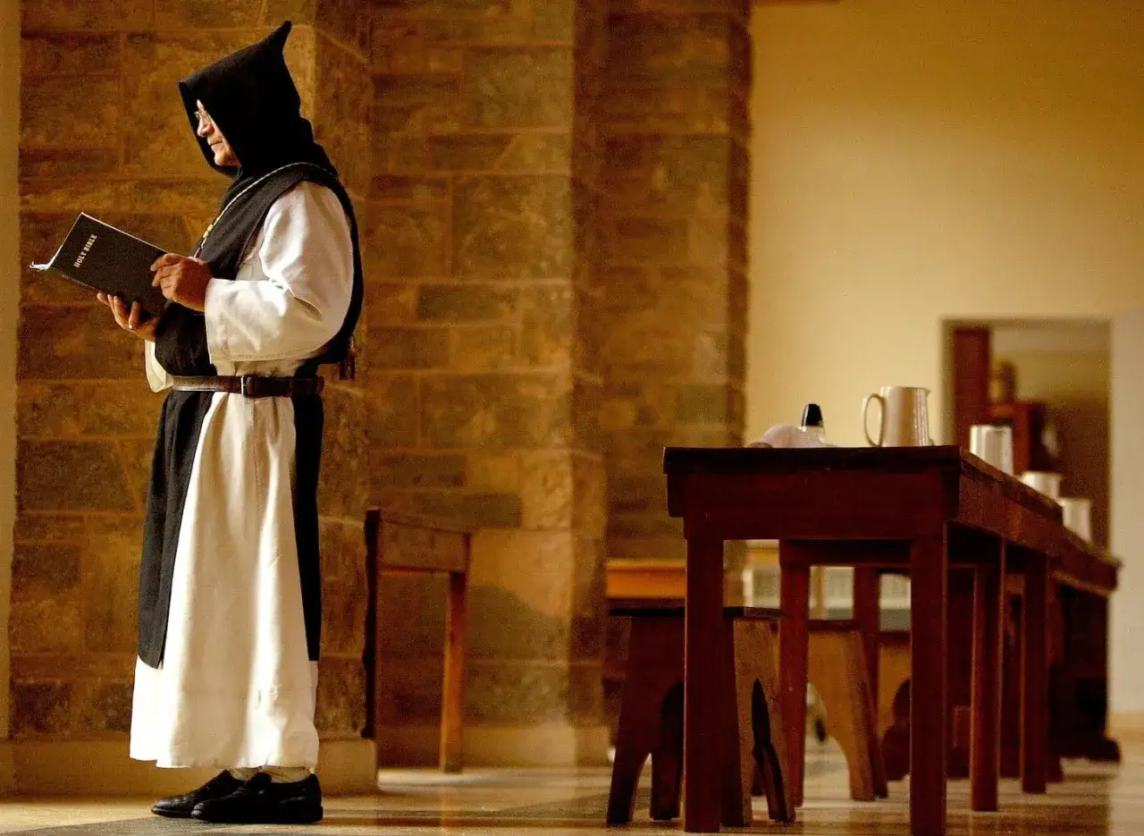 A monk in a black hooded robe and white tunic reads the Holy Bible. He stands beside a stone wall, a scene reminiscent of St. Benedict's teachings.