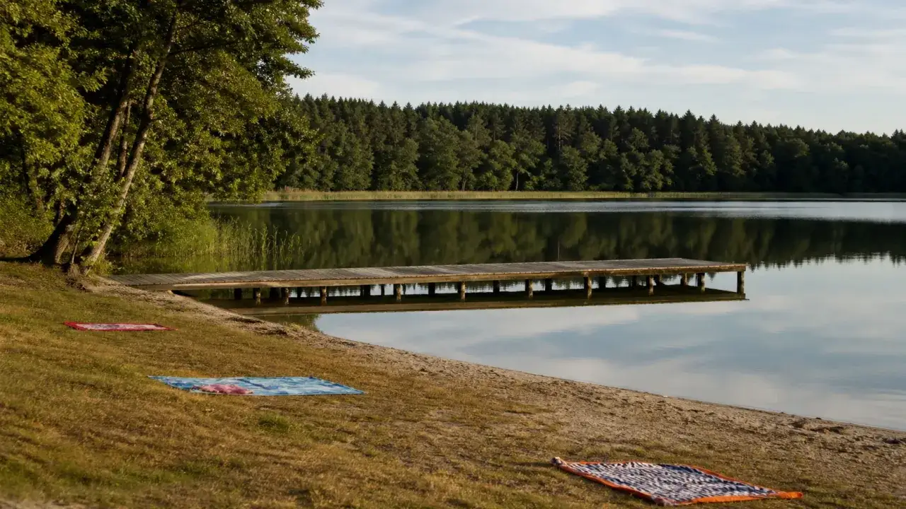 Drewniane molo nad spokojnym jeziorem, otoczonym lasem. Na trawie rozłożone koce, idealne na wypoczynek na polu biwakowym Salamonowo.