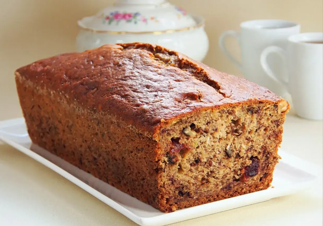Chlebek bananowy kwestia smaku, z orzechami i żurawiną, na białej paterze, w tle filiżanki i cukiernica.