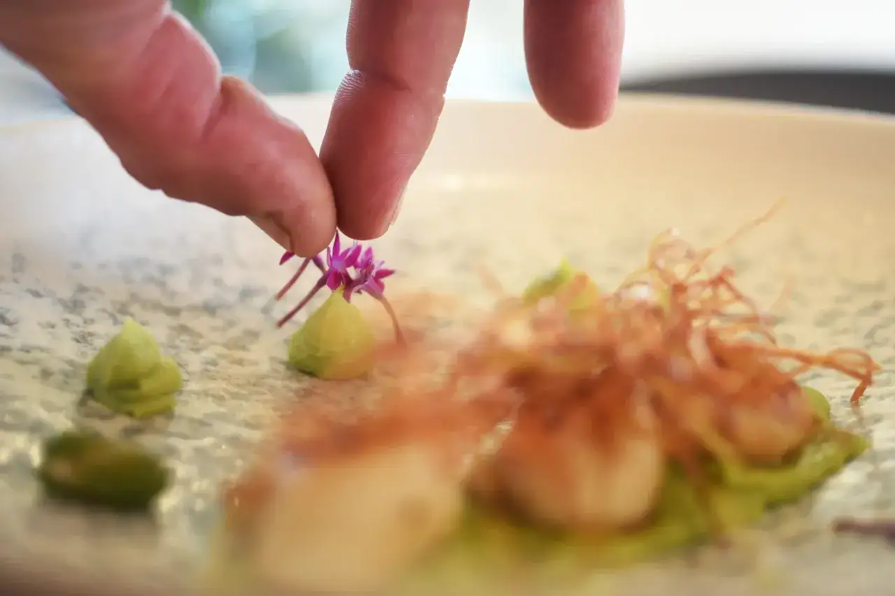 En hand dekorerar en tallrik med en liten lila blomma, en fin detalj f&ouml;r att ge **europa tips** om matlagning.