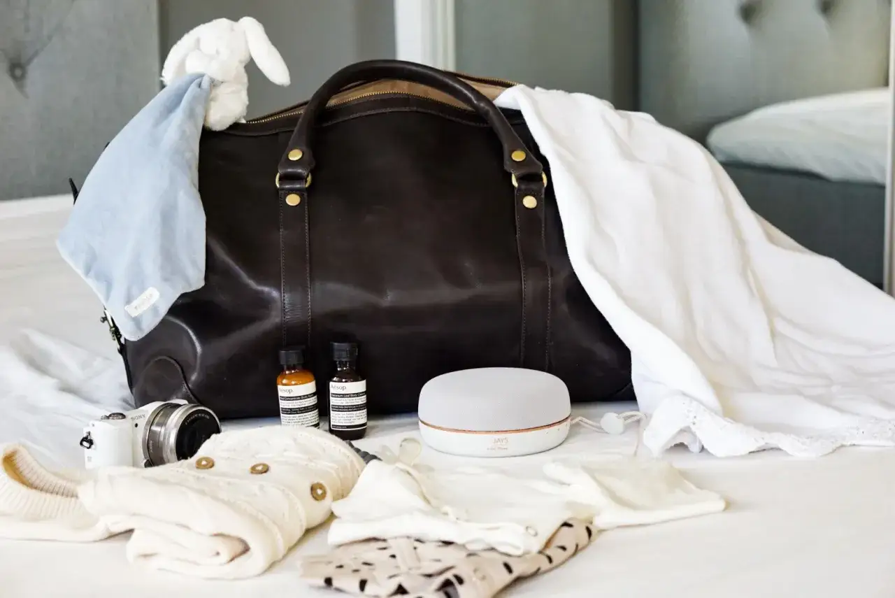 Packlista bb: m&ouml;rk l&auml;derv&auml;ska med bebiskl&auml;der, nalle, kamera, h&ouml;gtalare och Aesop-flaskor.