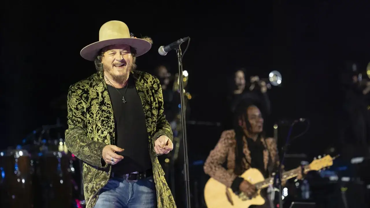 Un cantante con cappello e giacca damascata canta sul palco, accompagnato da musicisti. Un'atmosfera da concerto che evoca canzoni country famose.