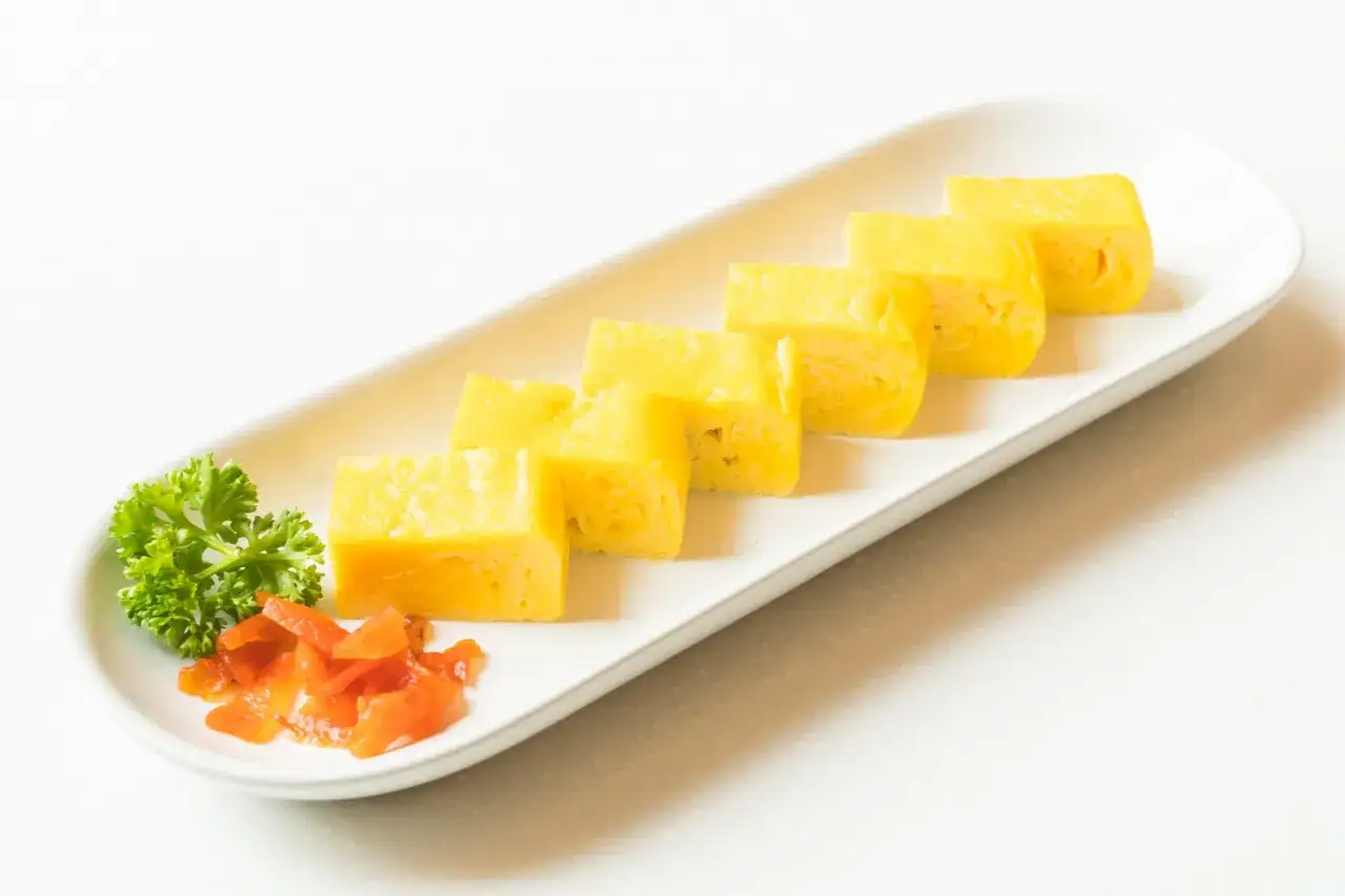Sześć kawałków tamago, czyli japońskiego omletu, ułożonych na białym talerzu. Obok gałązka pietruszki i pomarańczowe marynowane warzywa.