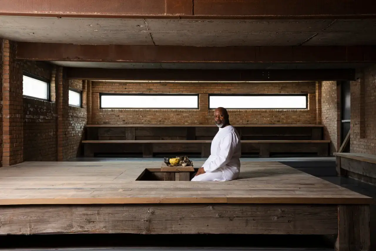 Theaster Gates studio, a man in white sits on a wooden platform in a raw, industrial space with brick walls and large windows.