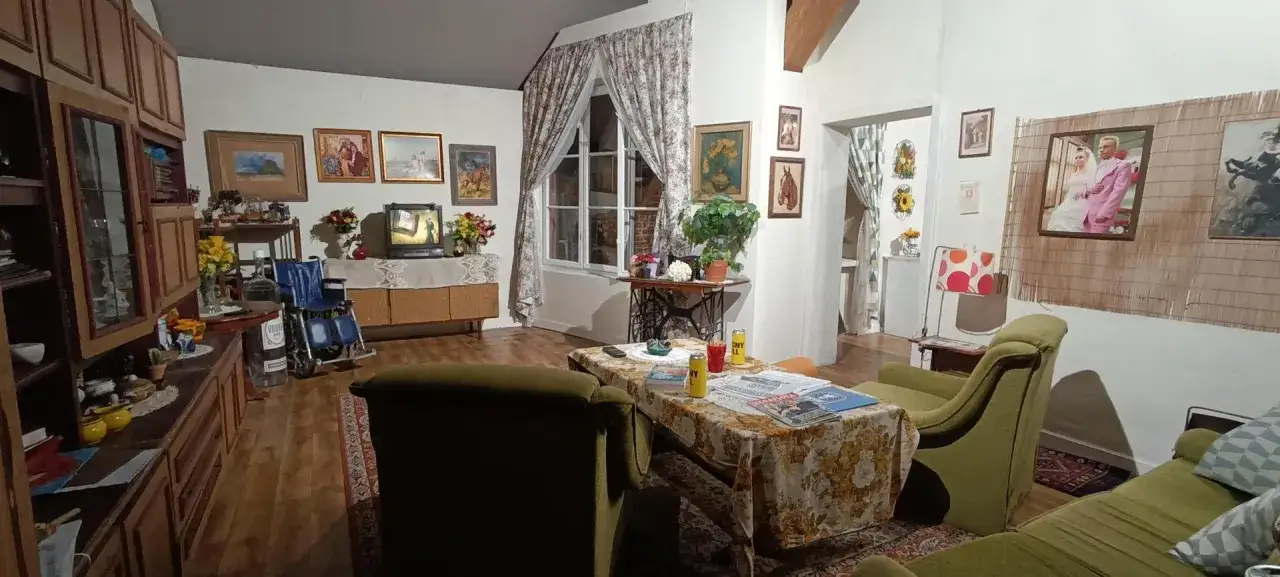Wnętrze mieszkania jak z **muzeum kiepskich**: kanapy, telewizor, wózek inwalidzki i zdjęcia na ścianach.