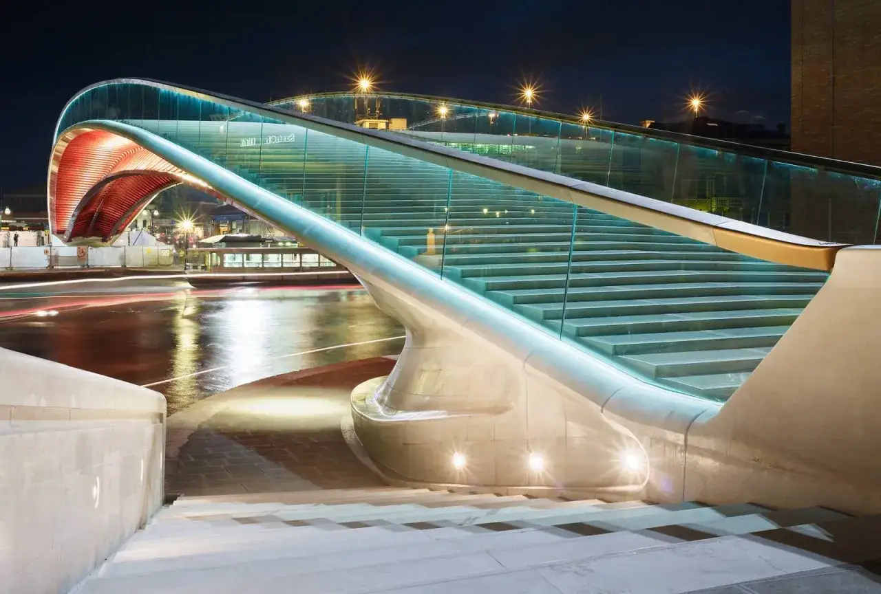Le **pont de la Constitution** s'illumine la nuit, ses escaliers de verre et de pierre menant vers un arc rouge audacieux au-dessus d'une rivi&egrave;re aux reflets lumineux.